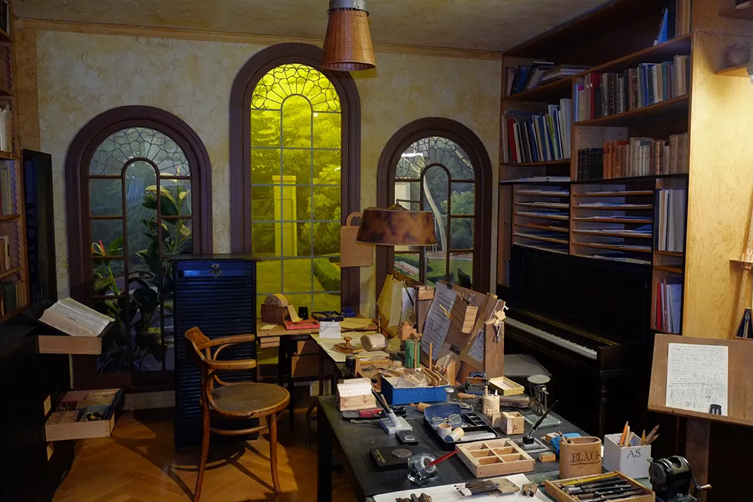 A replica of Arnold Schoenberg's study, with office furniture, an upright piano, and three large arched windows looking out on a garden